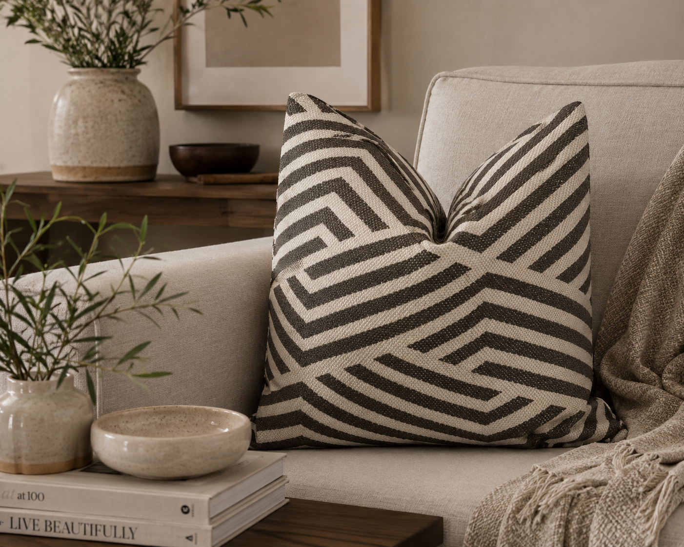 Coussin décoratif taupe et écru à motif chevron géométrique posé sur un canapé beige dans une ambiance chaleureuse et contemporaine avec décoration naturelle.