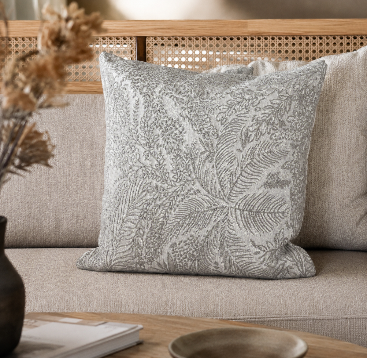 Coussin décoratif gris clair à motif feuillage dans une ambiance chaleureuse et naturelle avec canapé beige et décoration en bois.