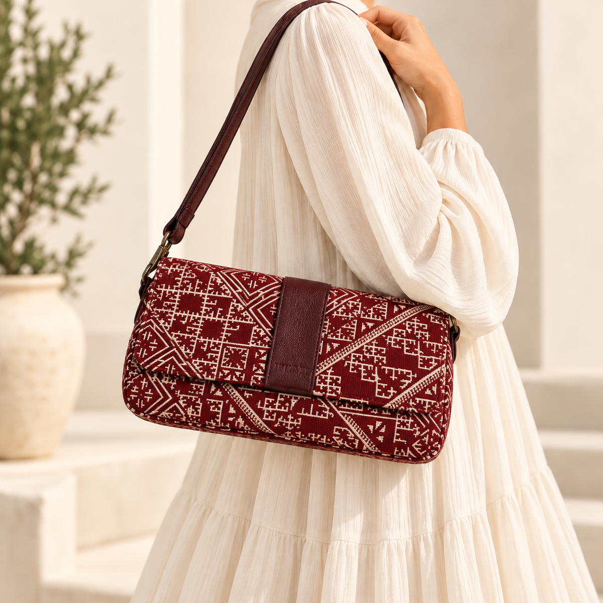 Sac baguette Biyadi Collection en tissu Tarz bordeaux à motifs marocains écrus et détails en cuir bordeaux, porté à l’épaule par une femme vêtue d’une robe longue et ample écrue.