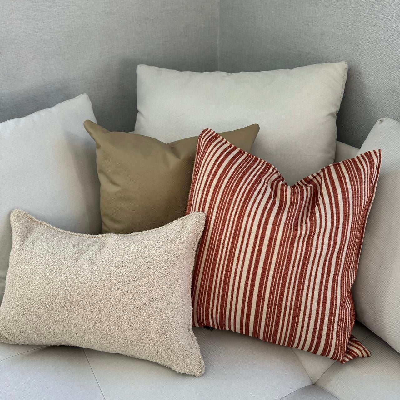 Composition de coussins décoratifs aux tons écrus et terracotta avec un coussin rayé rouge brique, un coussin bouclette écru et des coussins unis beige sur canapé moderne.