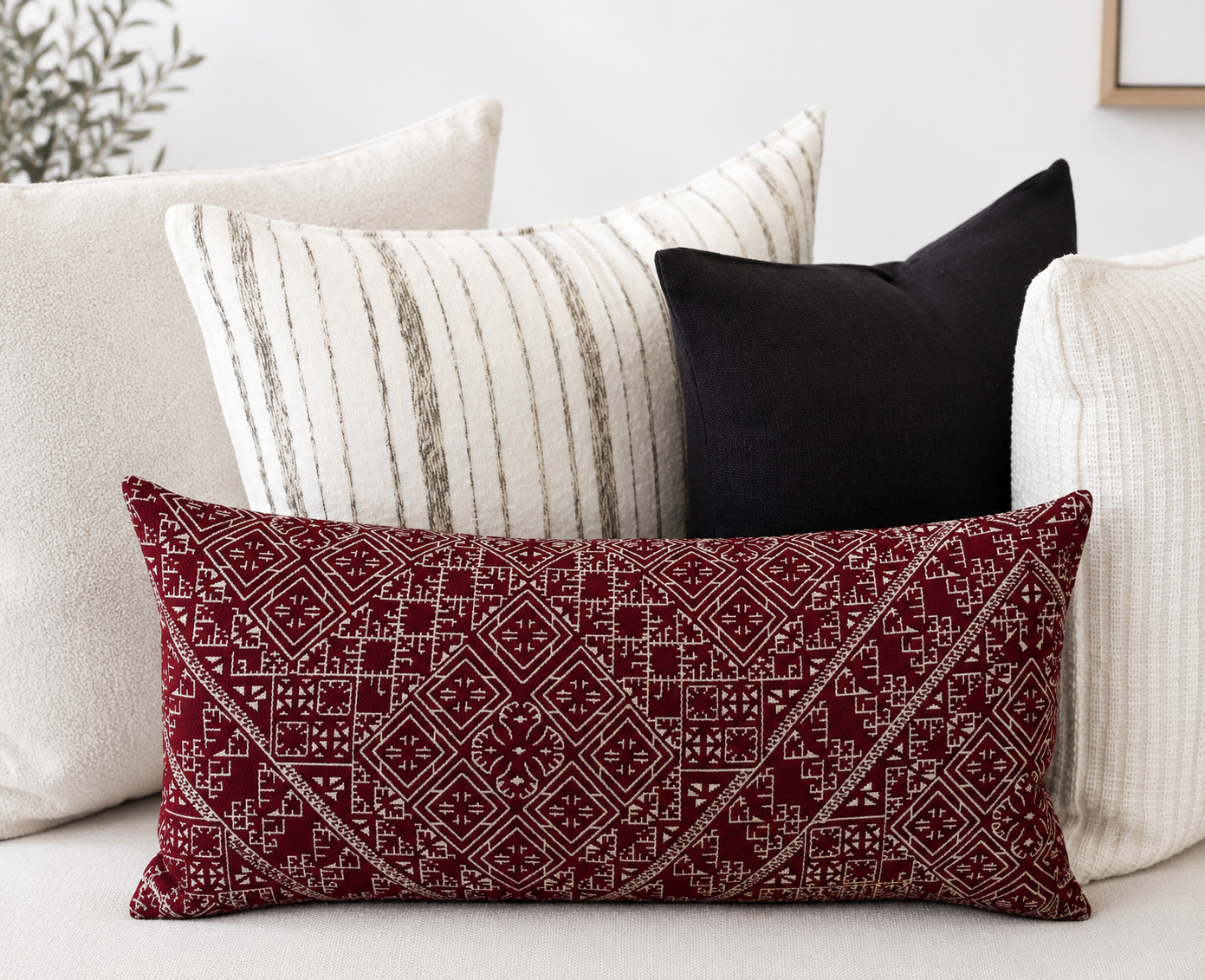 Composition de coussins décoratifs aux tons écru, noir et avec coussin marocain Tarz El Gharza bordeau à motif géométrique, ambiance élégante et contemporaine.