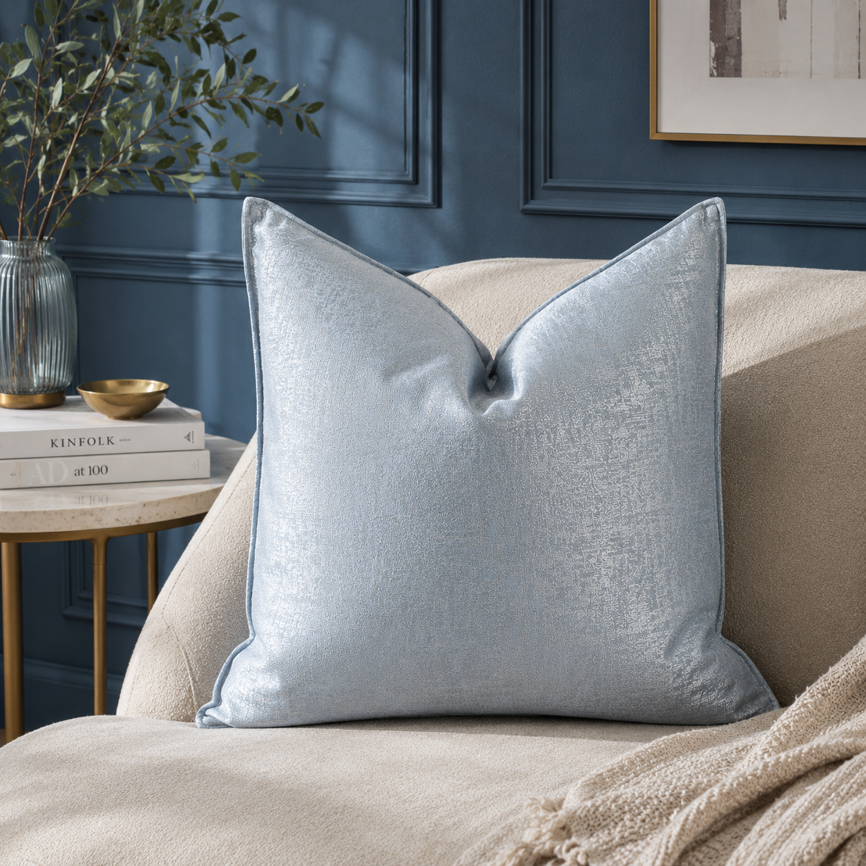 Coussin décoratif bleu glacé satiné posé sur un fauteuil beige dans une ambiance élégante avec mur bleu profond et décoration chic contemporaine.