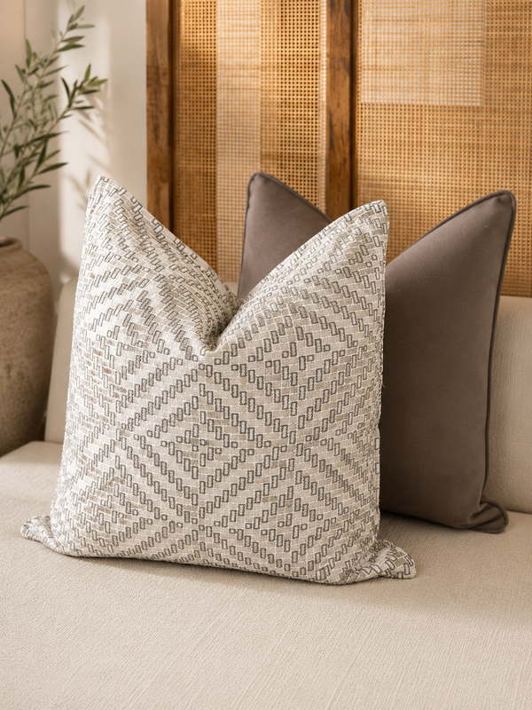 Coussin carré écru à motif géométriquet aupe en tissu tissé au Maroc, présenté avec un coussin taupe dans une ambiance de salon élégante aux tons naturels.