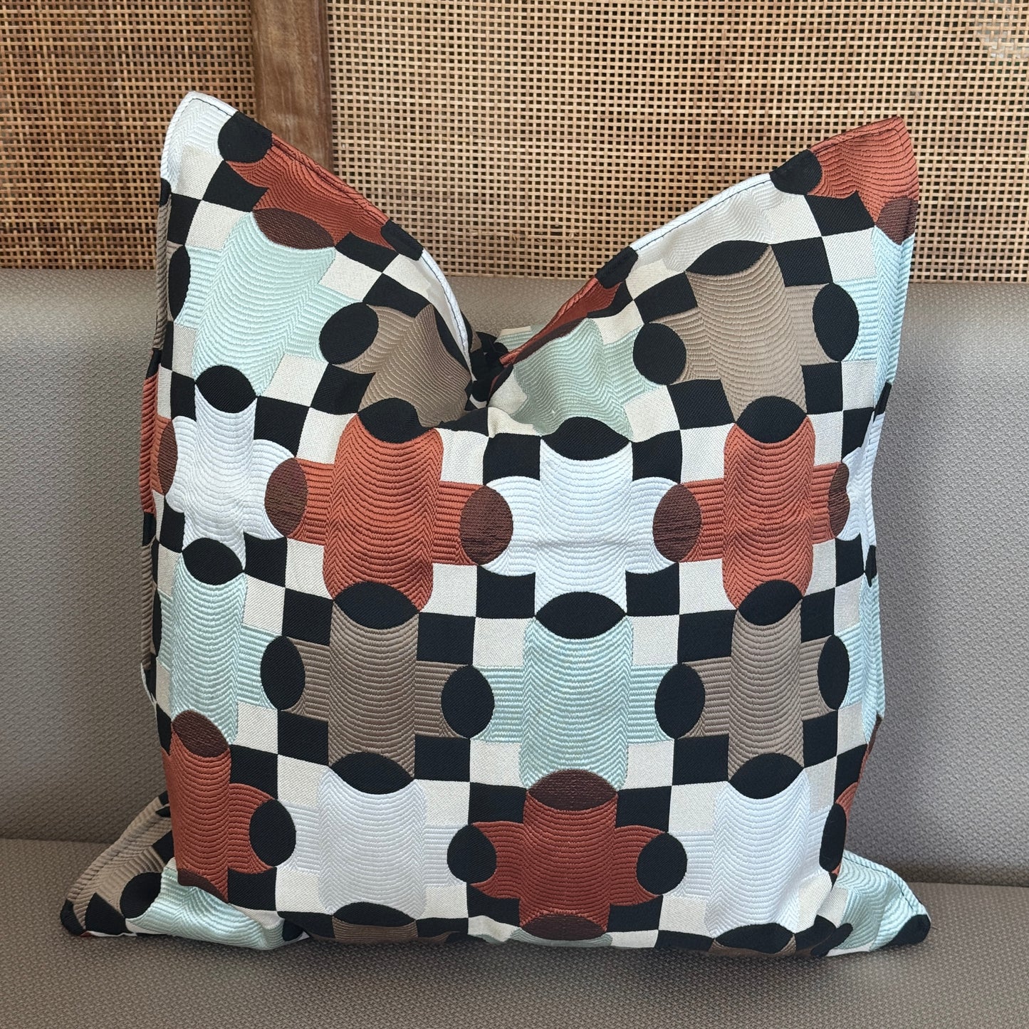 Coussin décoratif avec motifs géométriques colorés (orange, beige, taupe, noir et bleu clair) sur un canapé.

