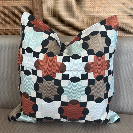 Coussin décoratif avec motifs géométriques colorés (orange, beige, taupe, noir et bleu clair) sur un canapé.

