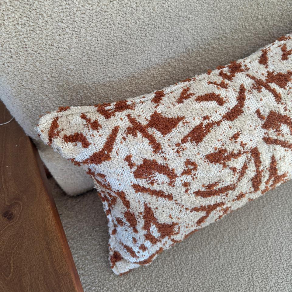 Détail d’un coussin rectangulaire en tissu texturé écru avec motif abstrait terracotta, finition soignée, posé sur fauteuil en bouclette beige