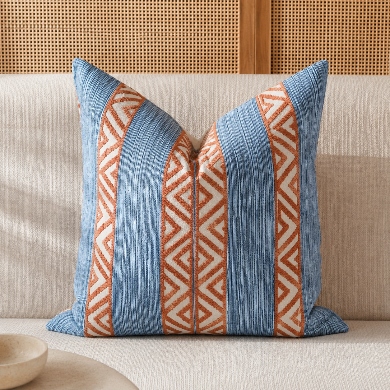 Coussin bleu et terracotta à motifs géométriques dans une ambiance néobeldi chaleureuse avec canapé beige et décoration naturelle – Biyadi Collection.