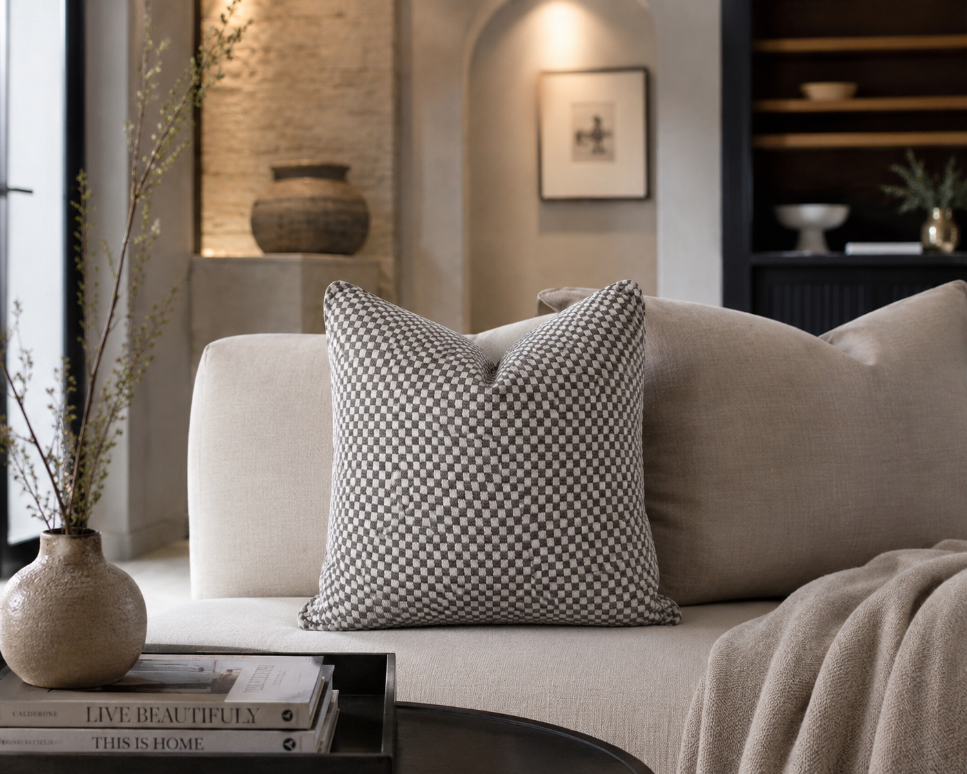 Coussin décoratif à petits carreaux gris et écru posé sur un canapé beige dans une ambiance intérieure élégante et minimaliste aux tons neutres.