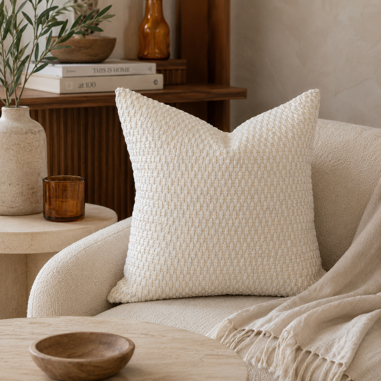 Coussin décoratif écru en tissu texturé tissé en relief, mis en scène dans un salon moderne aux tons naturels avec fauteuil bouclette, bois foncé et décoration élégante.