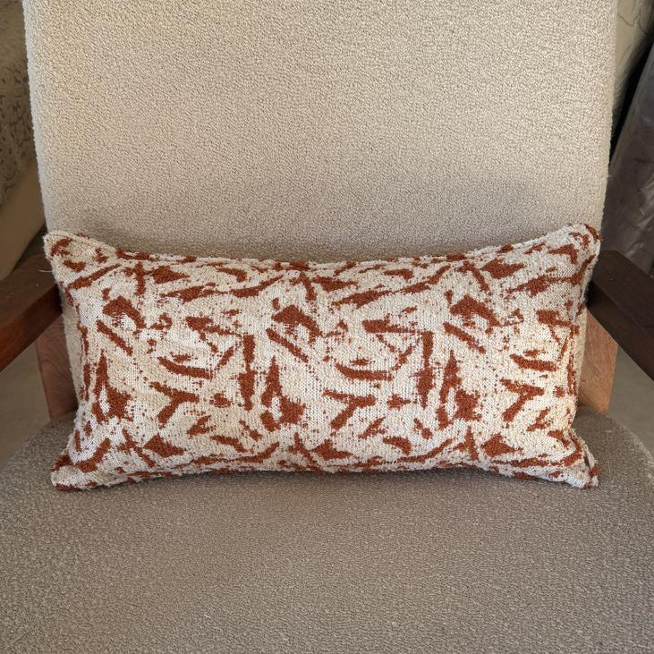 Coussin rectangulaire en tissu texturé écru avec motif abstrait terracotta, posé sur fauteuil en bouclette beige