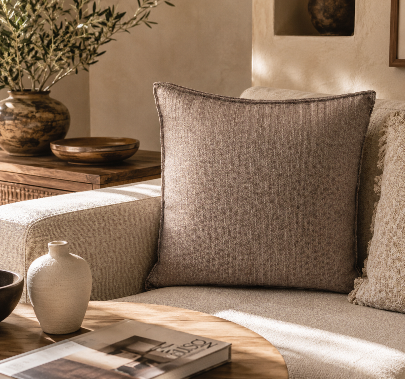 Coussin décoratif taupe à motif géométrique ton sur ton dans une ambiance chaleureuse et moderne, posé sur un canapé beige avec décoration élégante et naturelle.