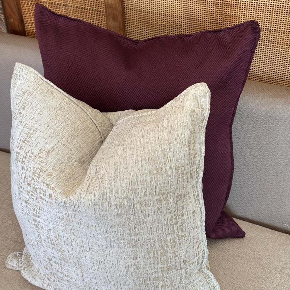 Deux coussins décoratifs superposés sur un canapé, un coussin uni burgundy à l’arrière et un coussin écru texturé à l’avant, style élégant et moderne.
