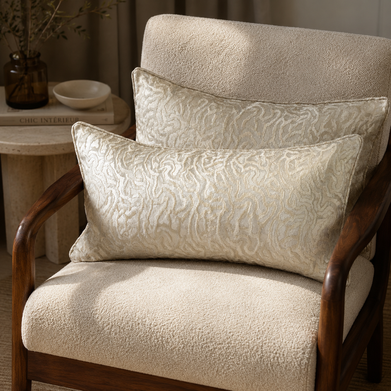 Deux coussins décoratifs beige clair à motif texturé en relief sur un fauteuil en bouclette écrue avec structure en bois foncé, ambiance élégante et chaleureuse.