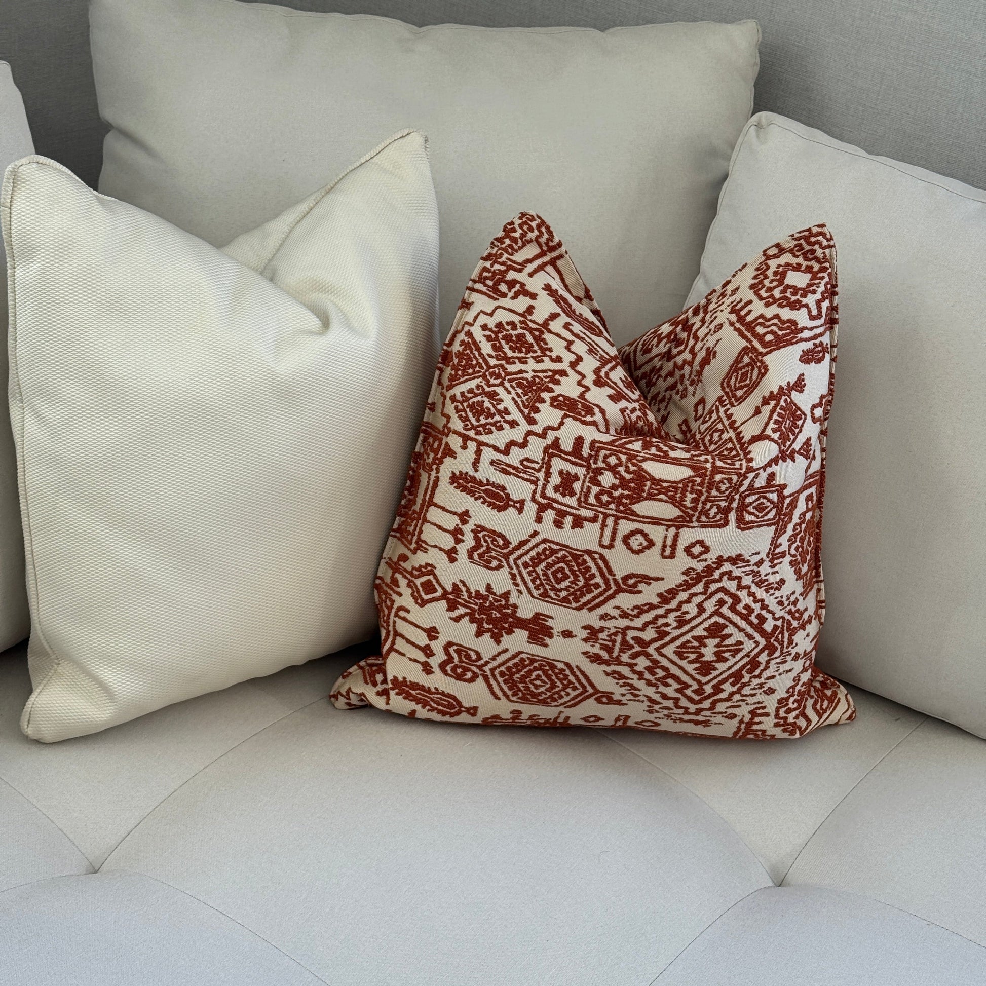 Composition de coussins décoratifs terracota et ecru à motifs ethniques et texture unie, présentés sur un fauteuil moderne avec une ambiance chaleureuse et élégante. Création Biyadi Collection.