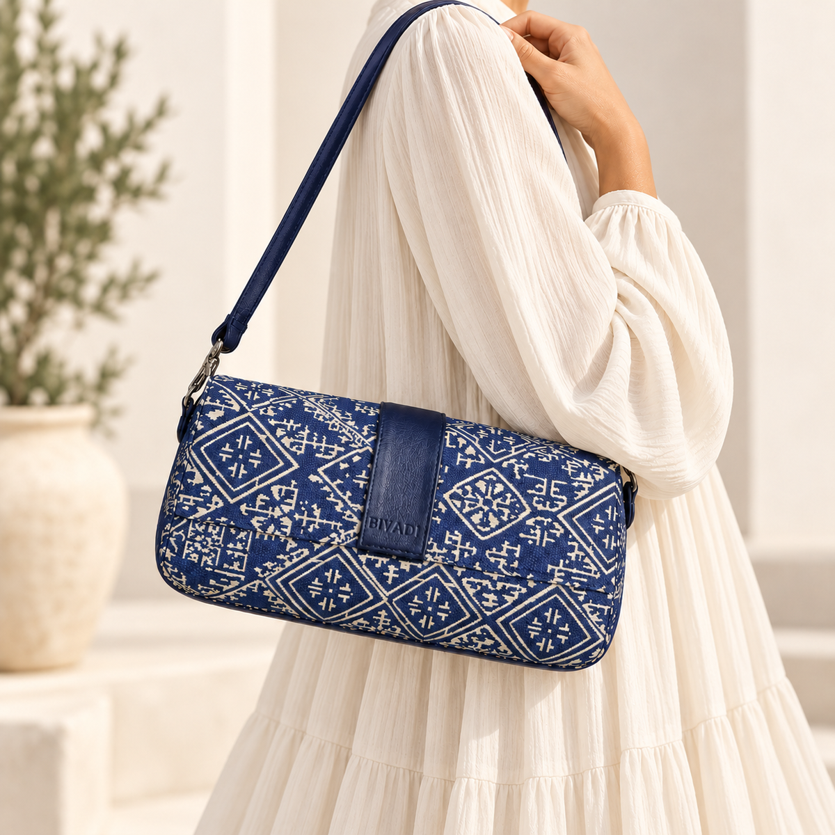 Sac baguette Biyadi Collection en tissu tarz bleu à motifs marocains écrus et détails en cuir bleu marine, porté à l’épaule par une femme vêtue d’une robe longue et ample écrue.