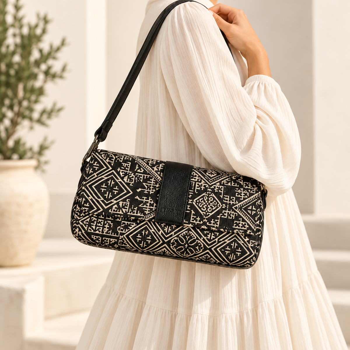Sac baguette Biyadi Collection en tissu Tarz noir à motifs marocains écrus, avec détails en cuir noir, porté à l’épaule par une femme vêtue d’une robe longue et ample écrue.