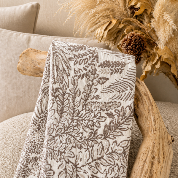 Tissu d’ameublement à motif feuillage taupe et écru tissé au Maroc, présenté dans une ambiance naturelle avec bois flotté et fleurs séchées