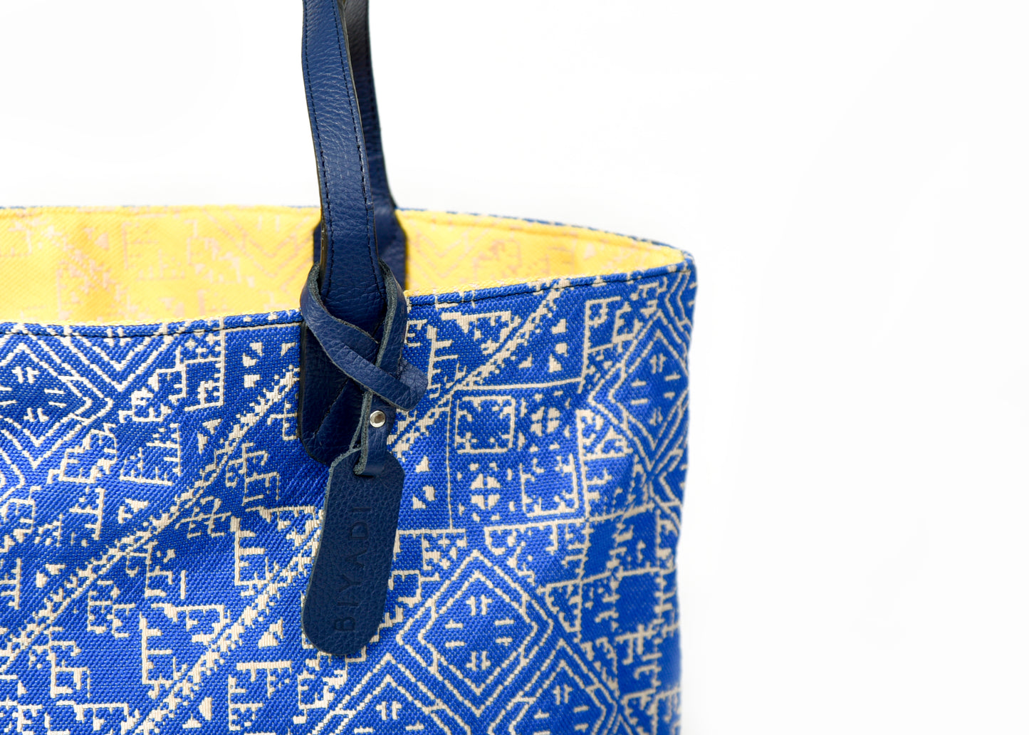 Détail du sac cabas reversible bleu et jaune en tissu marocain tissé avec anse en cuir noir et étiquette en cuir Biyadi Collection.
